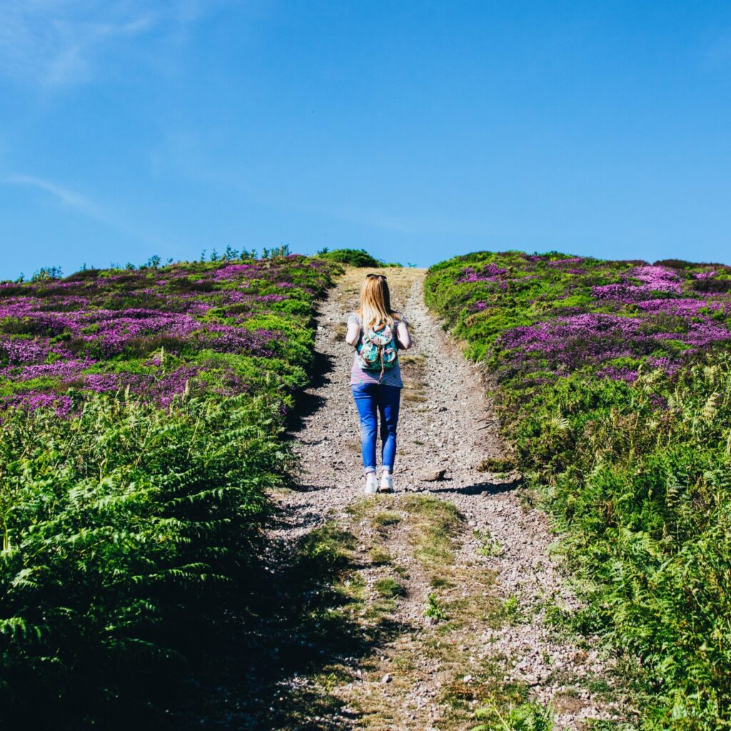Femme qui porte un sac à dos et qui est sur un chemin avec des fleurs violette